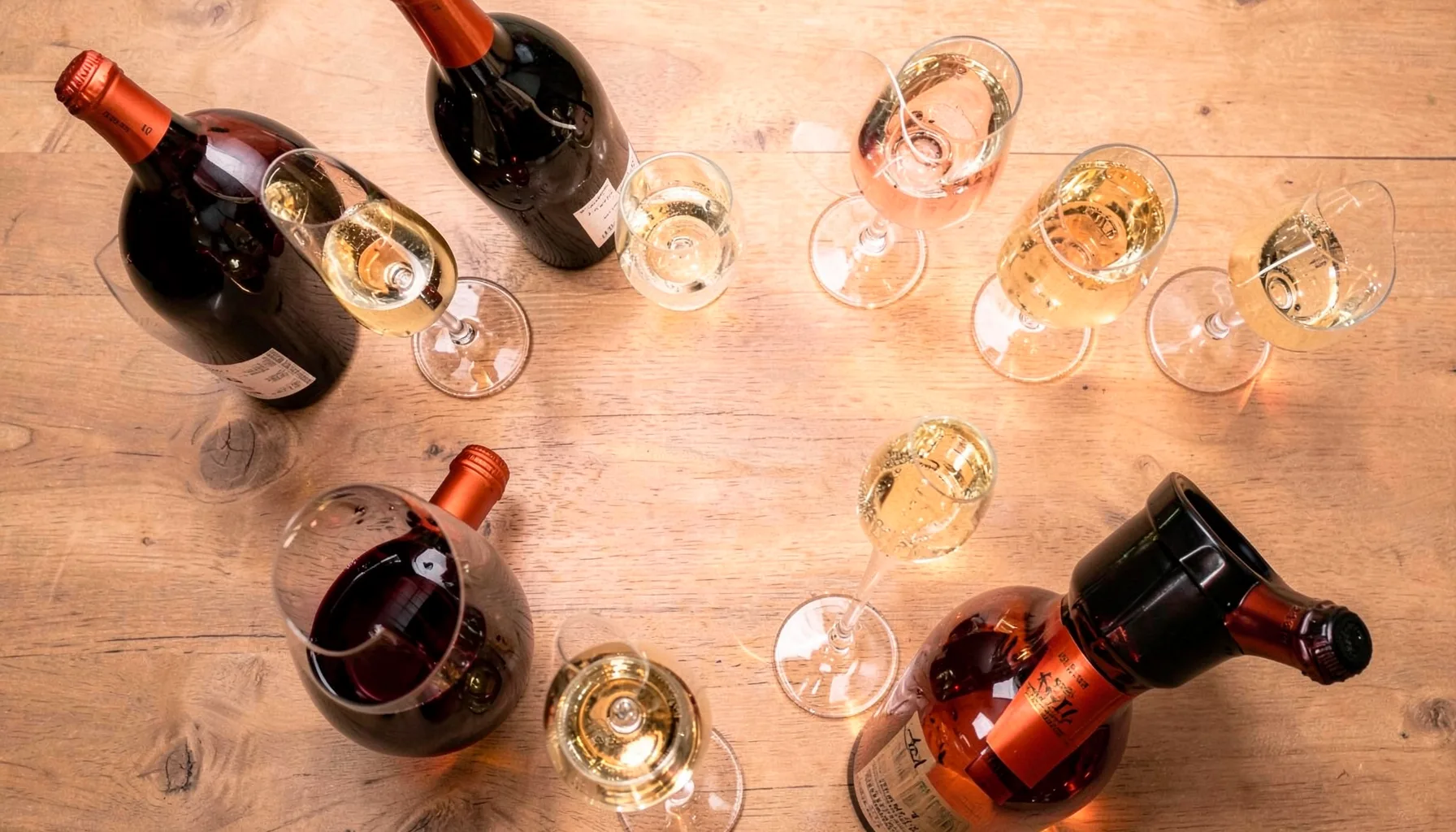Một bức ảnh chụp từ trên cao với những chai rượu vang đỏ và champagne đặt trên mặt bàn gỗ mộc, xung quanh là ly thủy tinh